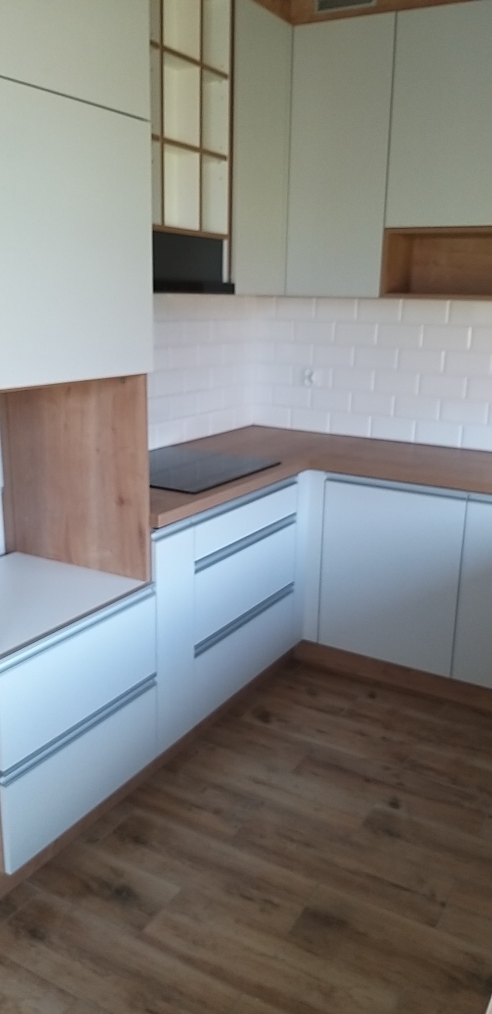 Nowoczesna kuchnia z białymi szafkami z poziomymi uchwytami, drewnianym blatem i panelami oraz białymi płytkami typu metro na ścianie, widok z perspektywy podłogi z drewnianymi panelami.