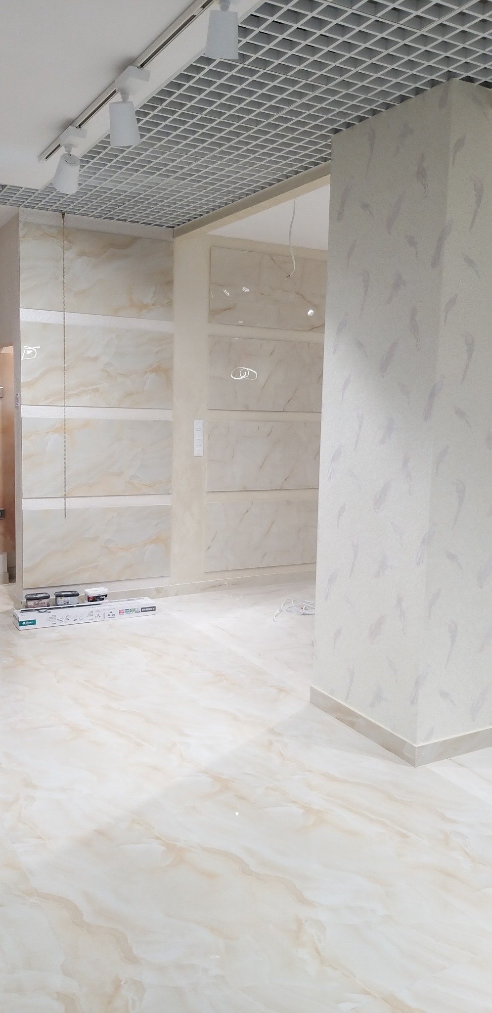 Wnętrze pomieszczenia z wykończonymi ścianami w płytkach imitujących marmur i wzorzystą tapetą na filarze, widoczne puszki z farbą i listwy przypodłogowe.