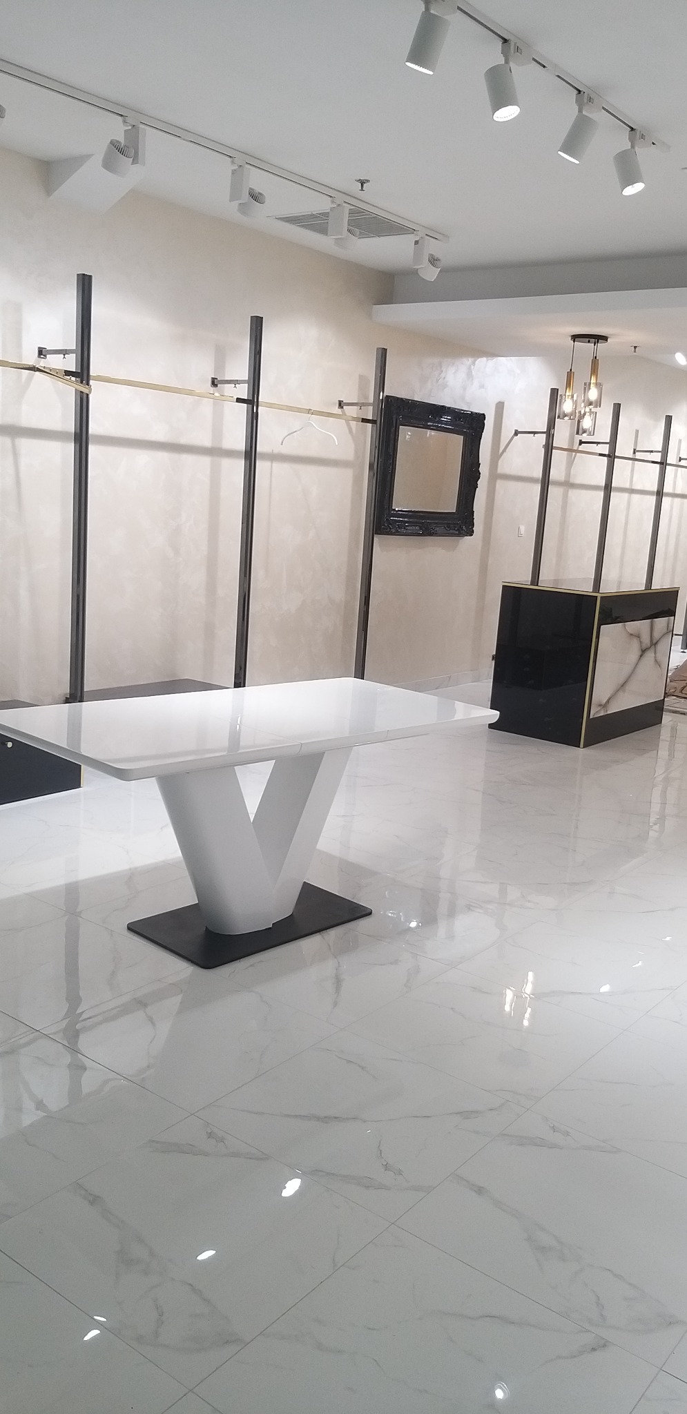 Błyszcząca, biała podłoga z marmurowym wzorem, nowoczesny biały stół z geometryczną podstawą na ciemnej stopie, w tle czarne metalowe wieszaki na ubrania, lustro w ozdobnej czarnej ramie i lada...