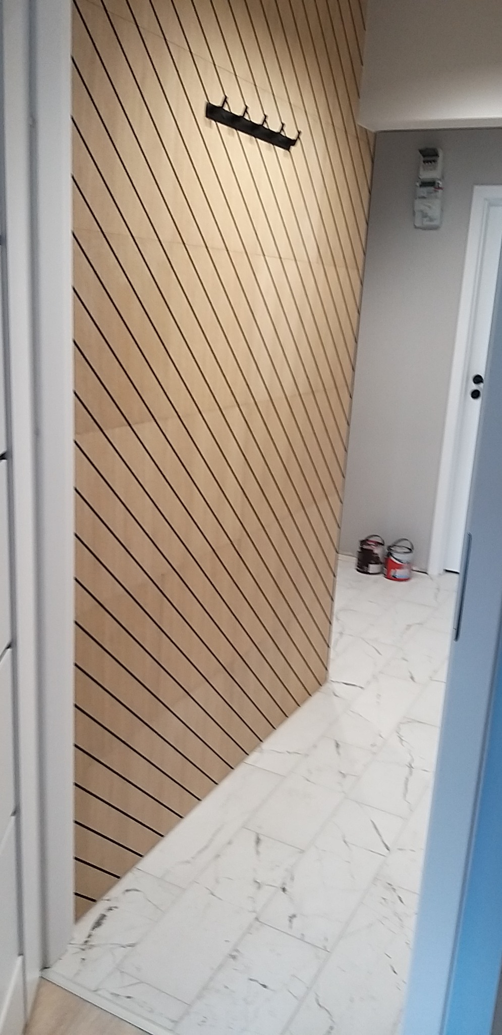 Nowoczesny przedpokój z drewnianą ścianą z ukośnymi listwami, wieszakiem na ubrania i podłogą z płytek imitujących marmur, widoczne puszki z farbą w tle.