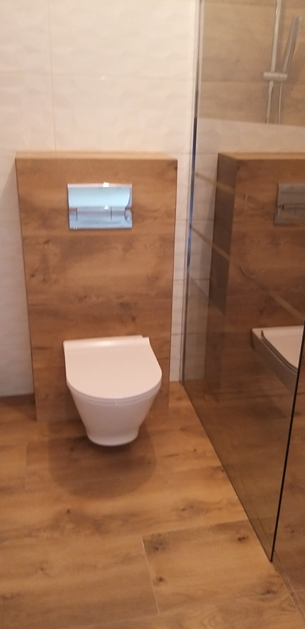 Nowoczesna toaleta podwieszana z panelem drewnopodobnym i chromowanym przyciskiem spłukującym, kabina prysznicowa ze szklaną ścianką w tle, podłoga z paneli imitujących drewno.