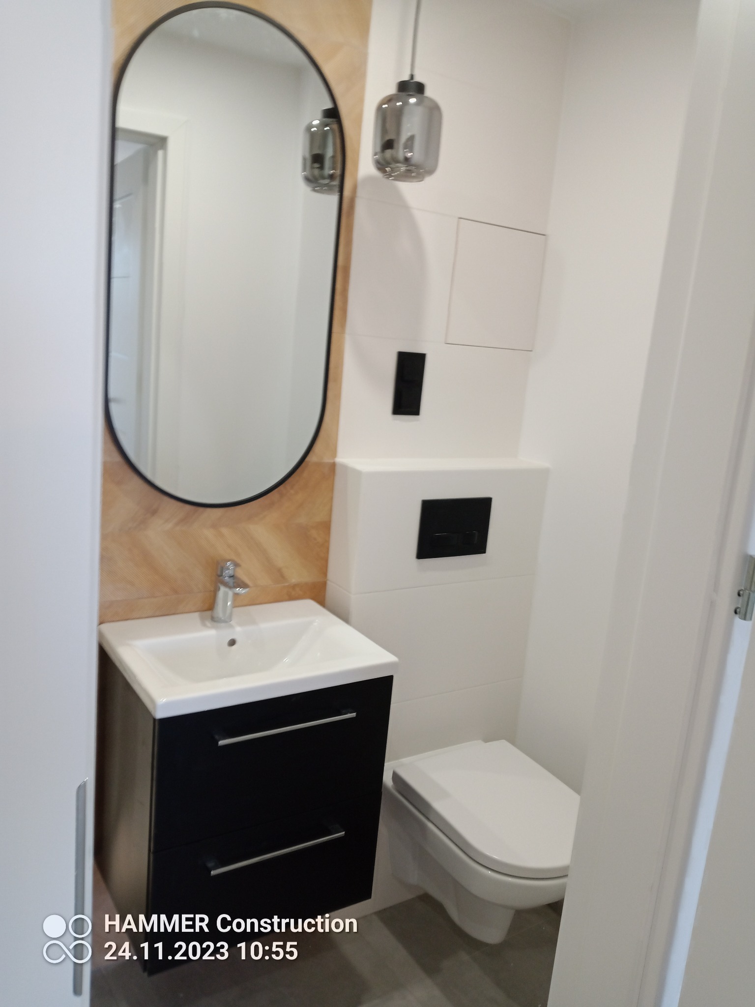 Nowoczesna łazienka po remoncie: czarna szafka pod umywalką z owalnym lustrem i drewnianą mozaiką w tle, wisząca toaleta, minimalistyczna lampa sufitowa.