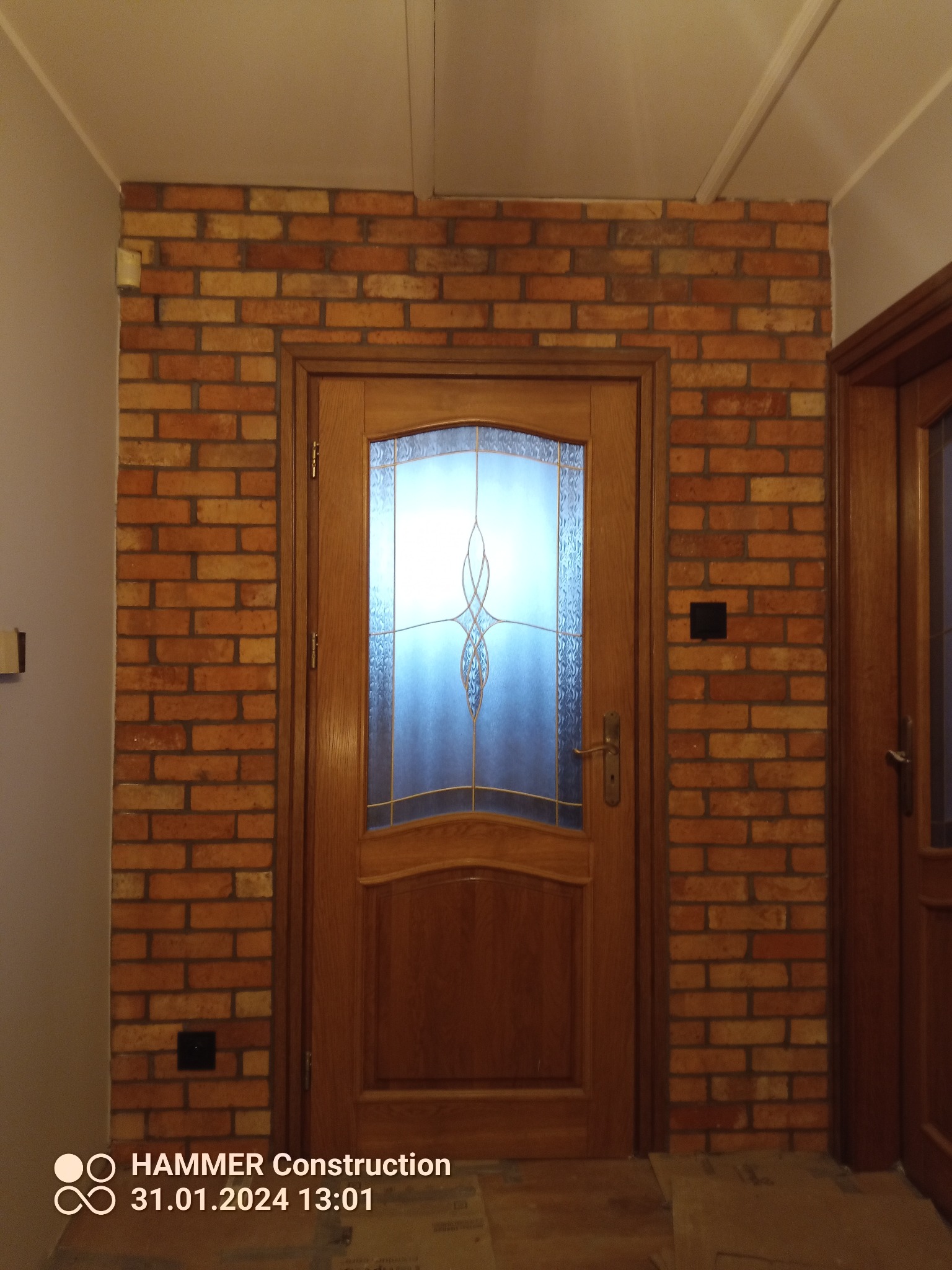 Wykończone wejście z drewnianymi drzwiami z szybą o ozdobnym wzorze, otoczone ścianą z cegły klinkierowej w odcieniach czerwieni i pomarańczy, z widocznym włącznikiem światła po prawej stronie.