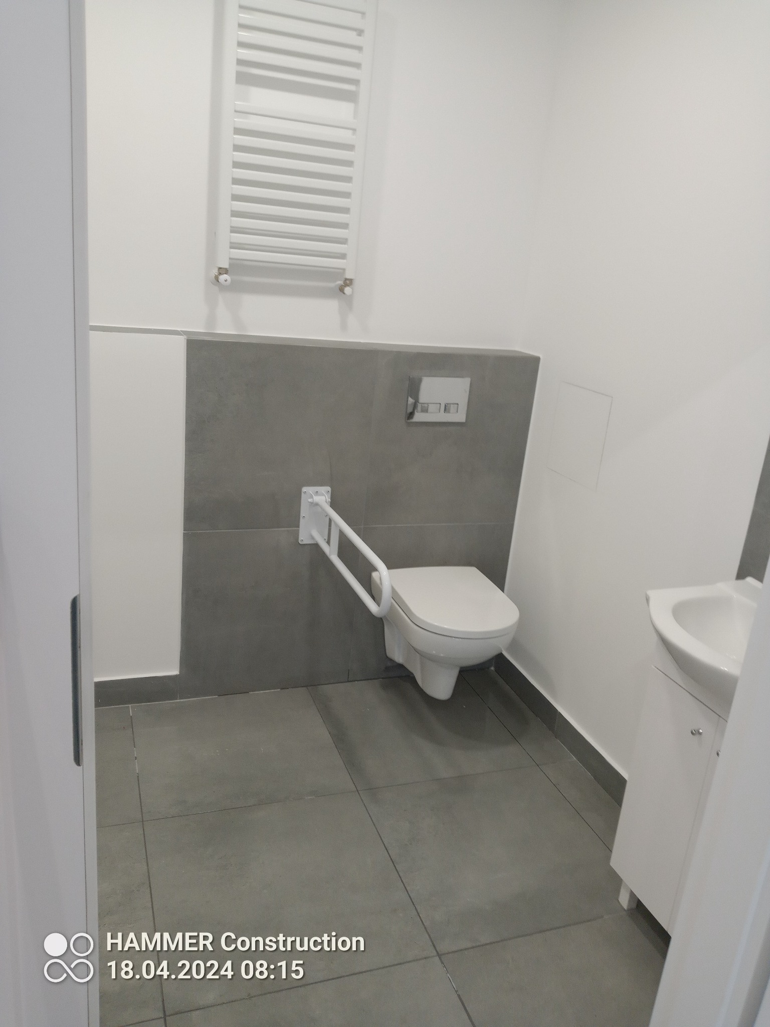 Nowoczesna, jasna łazienka z szarymi akcentami, wyposażona w toaletę z uchwytem dla osób z niepełnosprawnościami, białą umywalkę z szafką i grzejnik drabinkowy na ścianie.