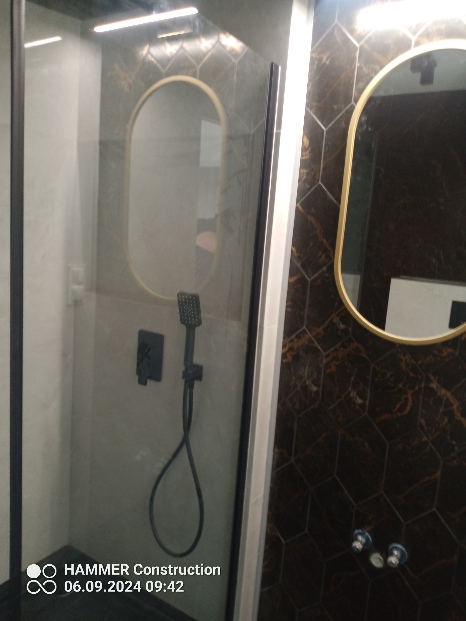 Nowoczesna kabina prysznicowa z deszczownicą i czarną armaturą na tle heksagonalnych płytek w odcieniach brązu i złota, uzupełniona owalnym lustrem z ramą w kolorze złota.