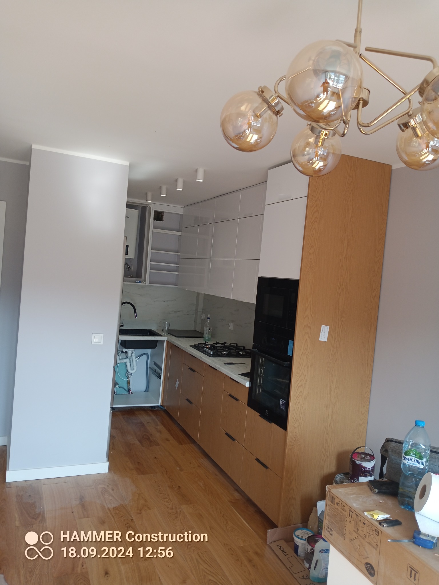 Nowoczesna kuchnia z drewnianymi i białymi szafkami, czarnym zlewem i marmurowym backsplashiem; widoczne elementy instalacji pod zlewem, oświetlenie sufitowe i bałagan na podłodze obok szafek.