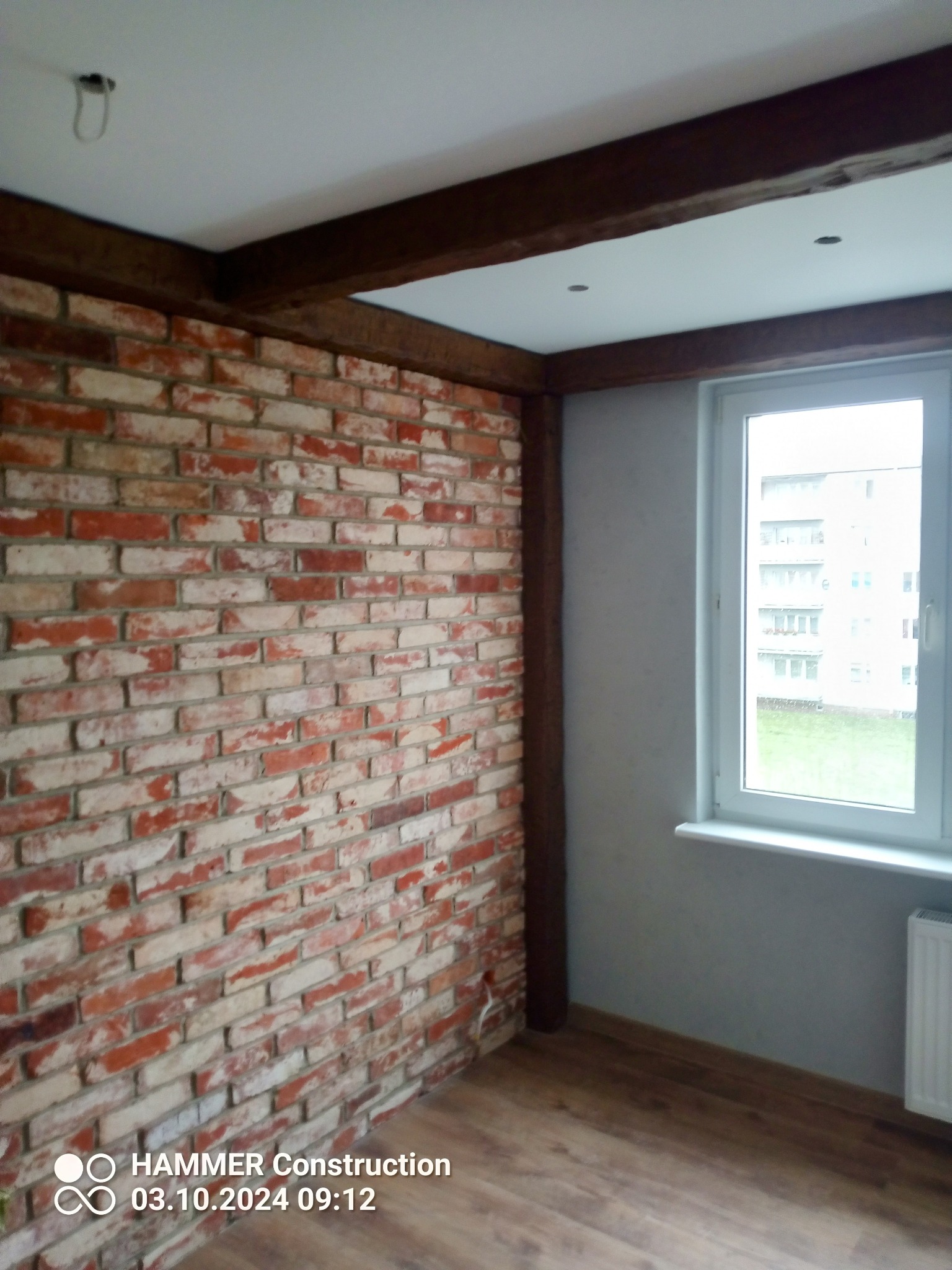 Ściana z czerwonej cegły z drewnianą ramą, jasne panele podłogowe i okno z widokiem na budynek mieszkalny.