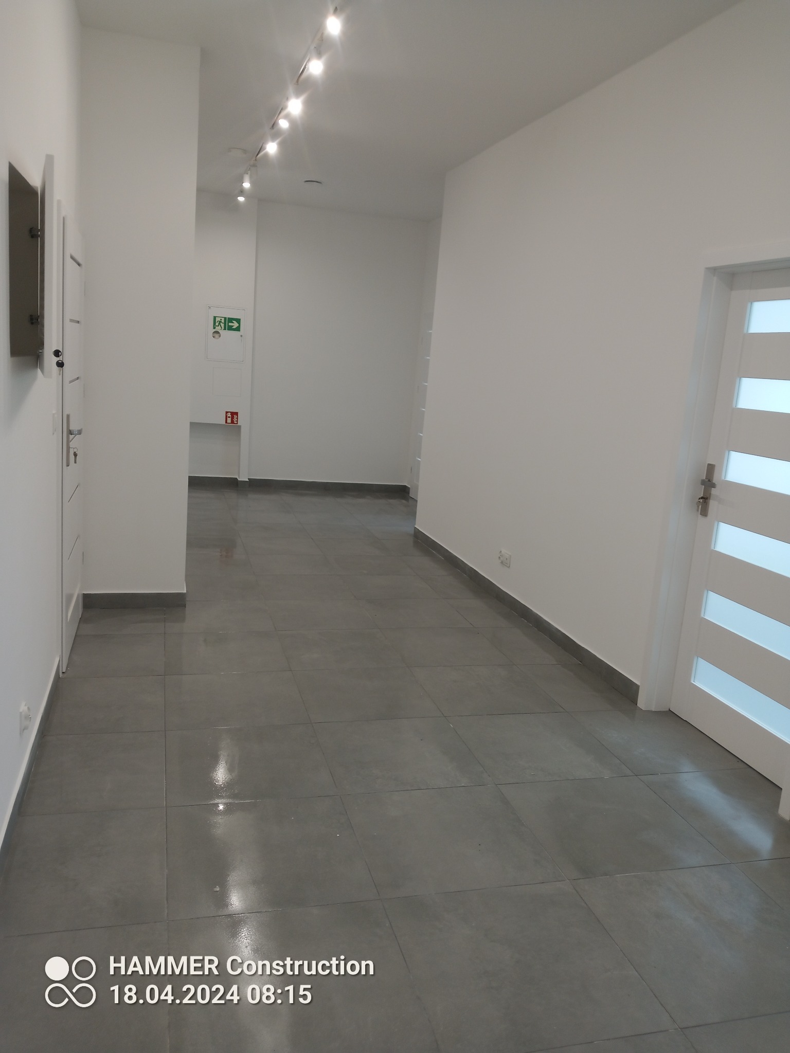Korytarz po remoncie z szarą, błyszczącą podłogą z płytek i białymi ścianami, widoczne oświetlenie sufitowe punktowe i drzwi z przeszkleniami.