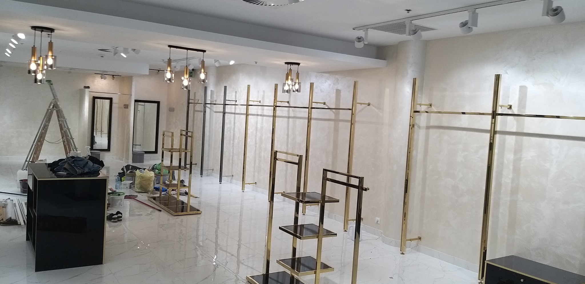 Wnętrze sklepu odzieżowego w trakcie aranżacji: złote regały na ubrania i półki, czarne konsole, drabina aluminiowa, widoczne ślady prac wykończeniowych na podłodze i ścianach.