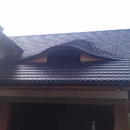 Ciemna dachówka w kształcie rybiej łuski kryje nowy dach domu w trakcie budowy. Widoczny fragment ściany z cegły i okno dachowe.