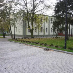 Szara kostka brukowa ułożona przed budynkiem z zielonym trawnikiem i drzewami. Widoczny właz kanalizacyjny i jesienne liście.