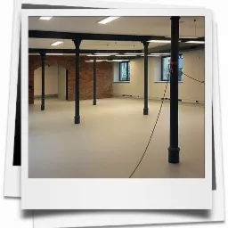 Wykończone wnętrze biurowe z kolumnami i ścianą z czerwonej cegły, oświetlone lampami sufitowymi, z widocznymi gniazdkami elektrycznymi wzdłuż ściany.