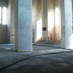 Wnętrze hali z betonowymi kolumnami w trakcie czyszczenia posadzki przez osoby w pomarańczowych kombinezonach, widoczny długi wąż i plakat na ścianie.