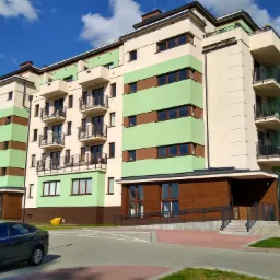 Nowoczesny budynek wielorodzinny z balkonami, otynkowany w odcieniach beżu i zieleni, z elementami drewnianymi na elewacji parteru, zaparkowany samochód na pierwszym planie.