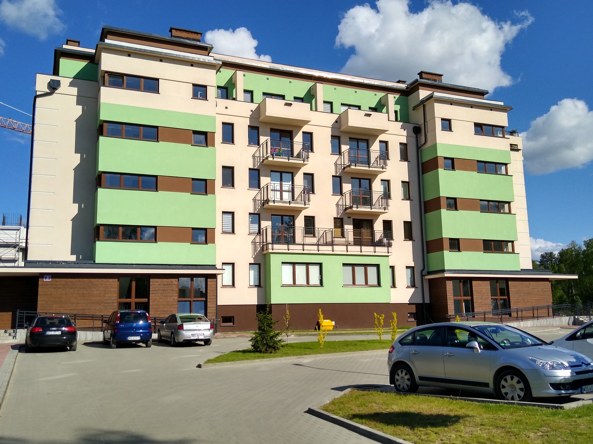 Nowoczesny budynek wielorodzinny z balkonami, pomalowany w pasy beżu, zieleni i brązu, z trzema samochodami zaparkowanymi przed nim w słoneczny dzień.