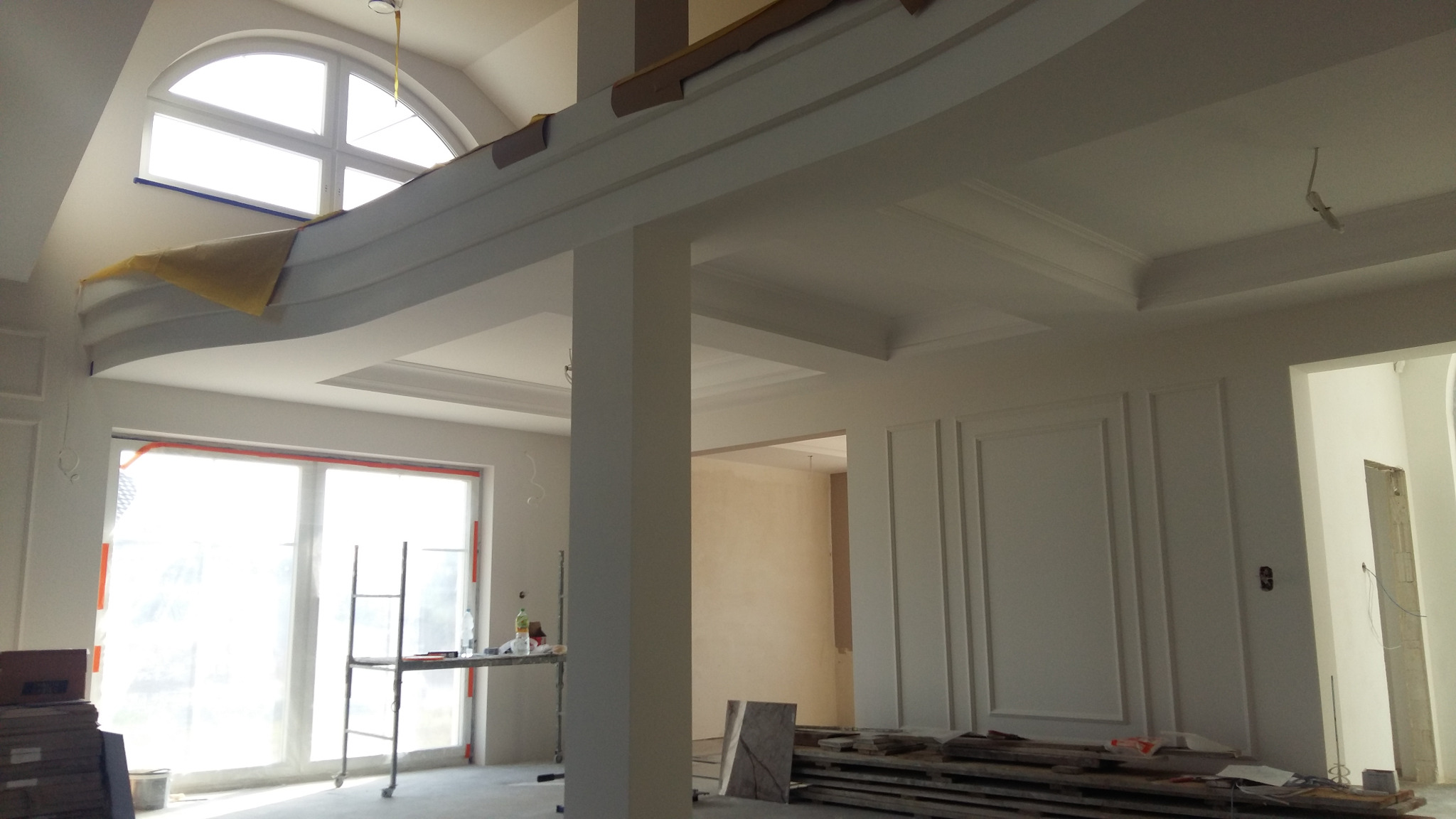 Wnętrze w trakcie prac wykończeniowych: widoczne sztukaterie na ścianach i suficie, okno łukowe z zabezpieczeniem folią, rusztowanie przy oknie, oraz przygotowane deski podłogowe.