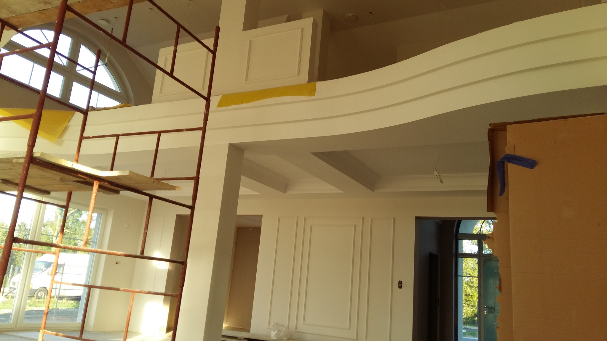 Wnętrze domu w trakcie wykańczania, widoczne rusztowanie, dekoracyjne listwy ścienne, sufit z elementami sztukaterii, okno łukowe i zabezpieczone folią elementy.