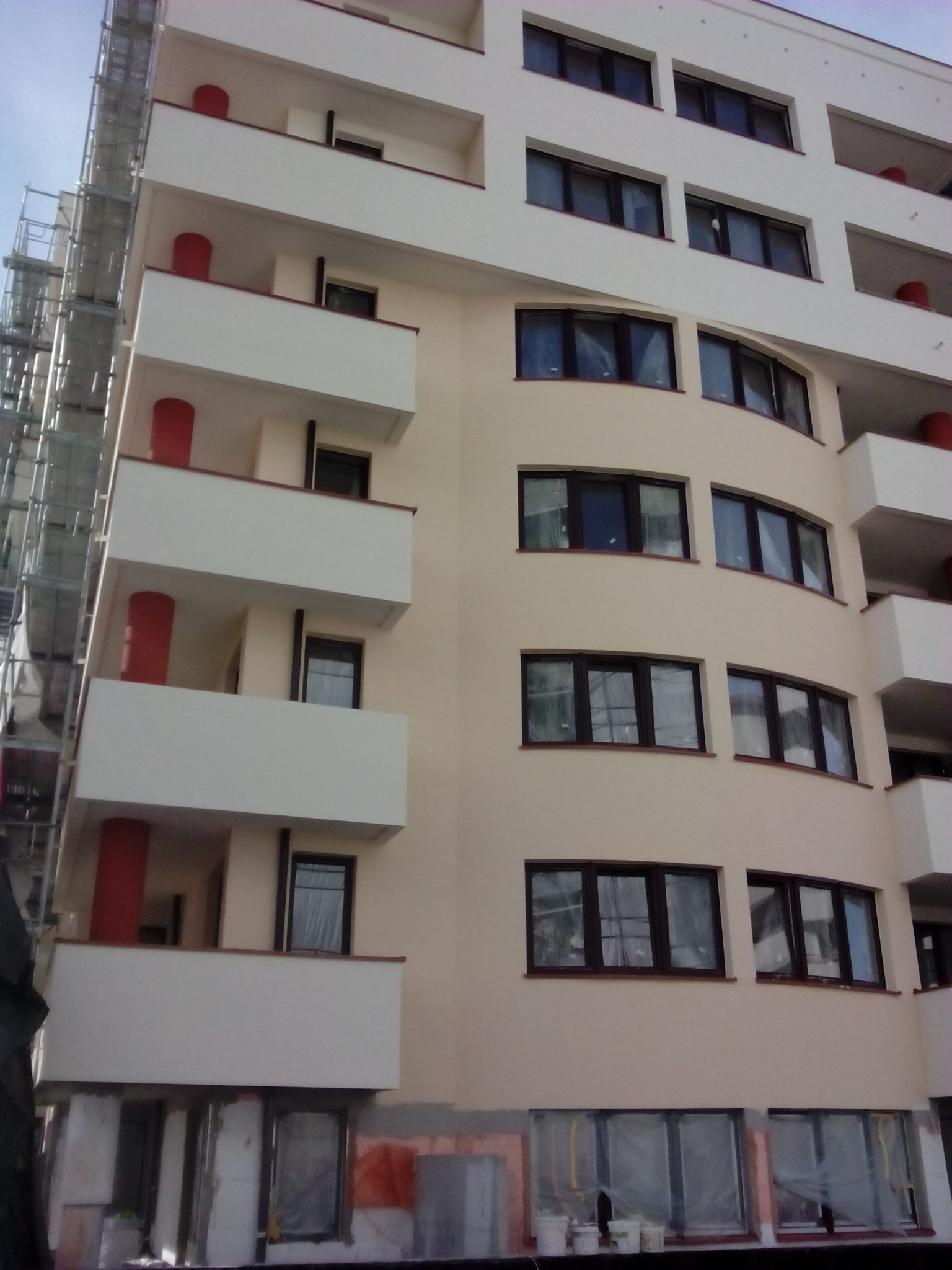 Fasada wielopiętrowego budynku z balkonami, częściowo pokryta rusztowaniem, okna zabezpieczone folią, widoczne wiadra z farbą sugerujące świeże malowanie.