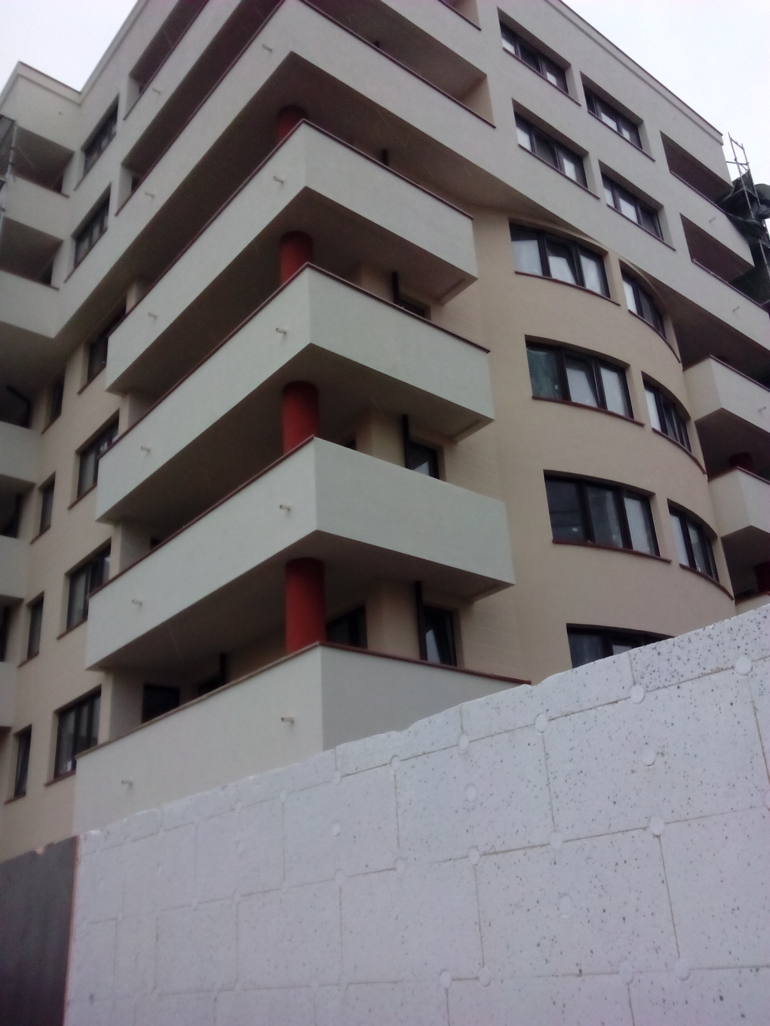 Wielopiętrowy budynek z balkonami o geometrycznym designie, widoczna ściana z białych płyt styropianowych w dolnej części kadru.