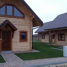 Dwa parterowe budynki z bali, z brązowymi ramami okiennymi i drzwiami, połączone ścieżką z kostki brukowej, na trawniku z szarą skrzynką elektryczną po prawej stronie.