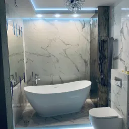 Nowoczesna łazienka z wolnostojącą białą wanną, marmurowymi płytkami na ścianach i podłodze, podświetleniem LED przy podłodze i suficie oraz ozdobnym żyrandolem kryształowym. Widoczne elementy...