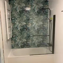 Nowoczesna łazienka z wanną obudowaną białymi płytkami, przeszklonym parawanem nawannowym z czarnymi elementami i ścianą wyłożoną dekoracyjnymi płytkami z motywem ptaków i roślin.