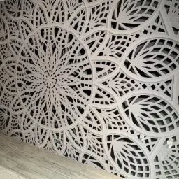Dekoracyjna ściana z ażurowym wzorem mandali w odcieniach bieli i szarości, widoczne gniazdka elektryczne, podłoga z paneli.