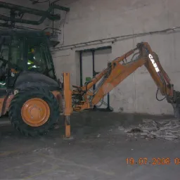 Pomarańczowa koparko-ładowarka CASE z młotem pneumatycznym kruszy betonową posadzkę wewnątrz hali. Operator w kabinie. Widoczne rury pod sufitem.