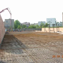 Siatka zbrojeniowa na budowie, widoczni pracownicy i pompa do betonu w tle. Częściowo wzniesione mury z czerwonej cegły.