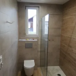 Nowoczesna łazienka z beżowymi płytkami, kabiną prysznicową ze szklanymi drzwiami i wiszącą toaletą. Płytki imitujące drewno w strefie prysznica. Widoczne narzędzia na p&oacute;łce pod oknem.