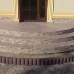 Wykończone kafelkami trójstopniowe schody wejściowe do budynku, z ozdobnym obramowaniem z brązowych palisad i nieregularną kamienną nawierzchnią u podstawy.