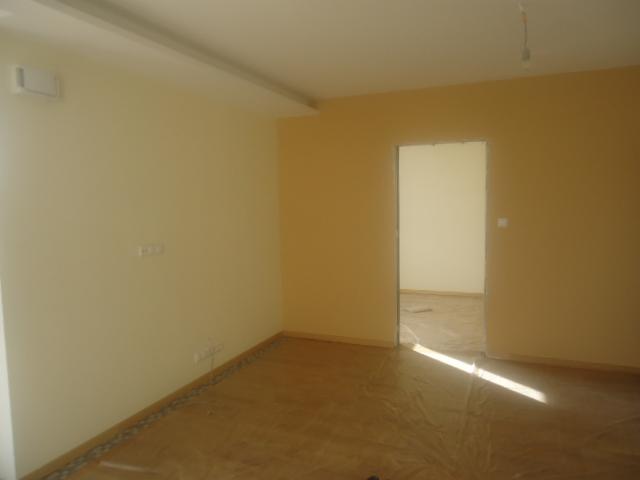 Pomalowane na dwa odcienie beżu puste pomieszczenie w trakcie prac wykończeniowych, podłoga zabezpieczona folią, widoczne gniazdka elektryczne i wiszący kabel od lampy.