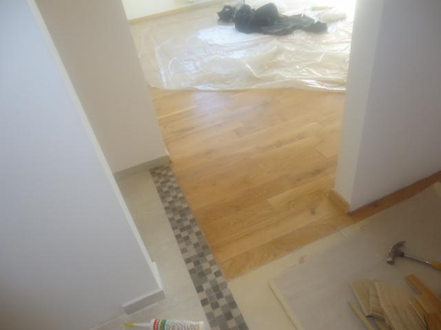 Układanie jasnej drewnianej podłogi w przejściu, widoczna mozaika na styku z innym pomieszczeniem, narzędzia i materiały budowlane w tle.