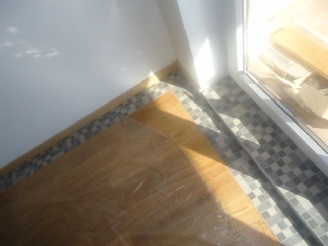 Fragment podłogi z przejściem z drewnianej powierzchni, zabezpieczonej folią, do mozaiki ceramicznej w odcieniach szarości przy oknie i ścianie.