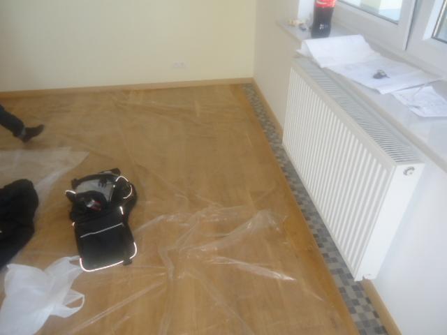Przygotowanie podłoża pod posadzkę: parkiet zabezpieczony folią malarską, widoczny grzejnik i torba narzędziowa.