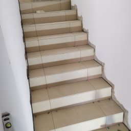 GUD-BUD - Schody w trakcie wykańczania, z jasnymi płytkami i widocznymi narzędziami glazurniczymi, poziomica przy ścianie, nierówne krawędzie ścian wymagające wykończenia.