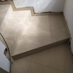 GUD-BUD - Wyłożone jasnymi płytkami schody w trakcie prac wykończeniowych, widoczne niedoskonałości na ścianach i suficie.