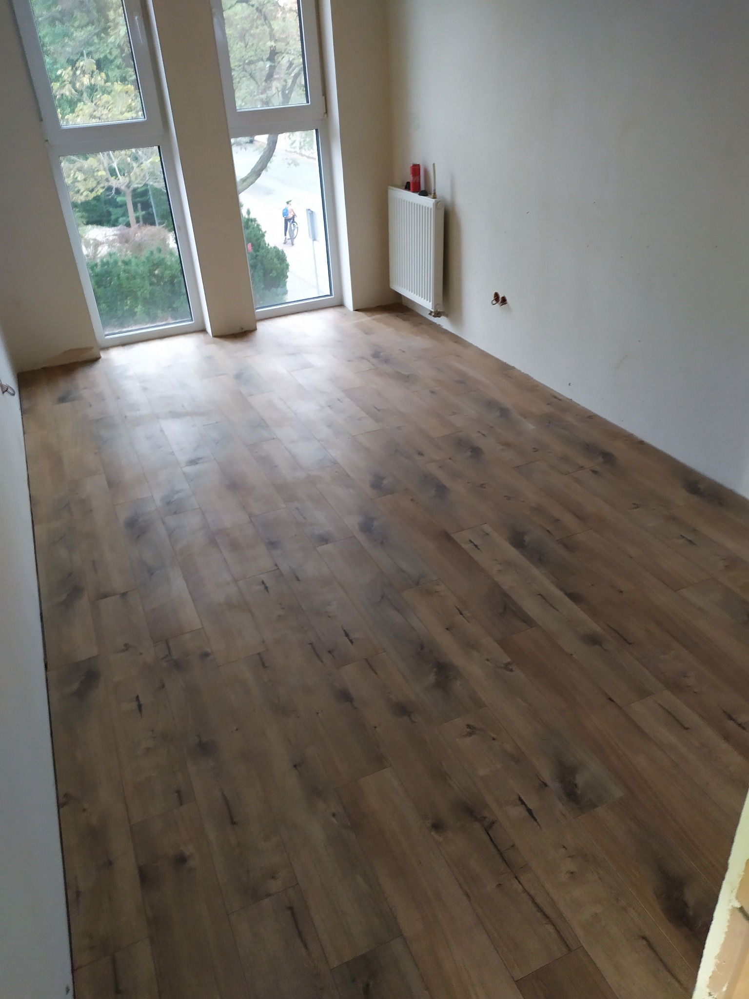 Ułożona podłoga z paneli imitujących drewno w pustym pokoju z oknem, widoczny fragment grzejnika i niedokończone wykończenie ścian.