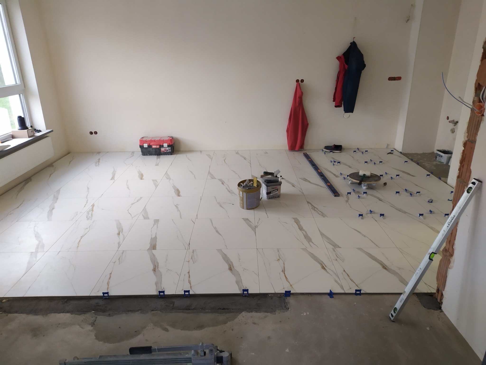 Układanie białych płytek imitujących marmur na podłodze w trakcie remontu, z użyciem poziomicy, krzyżyków dystansowych i narzędzi glazurniczych. W tle widoczna ściana i okno.
