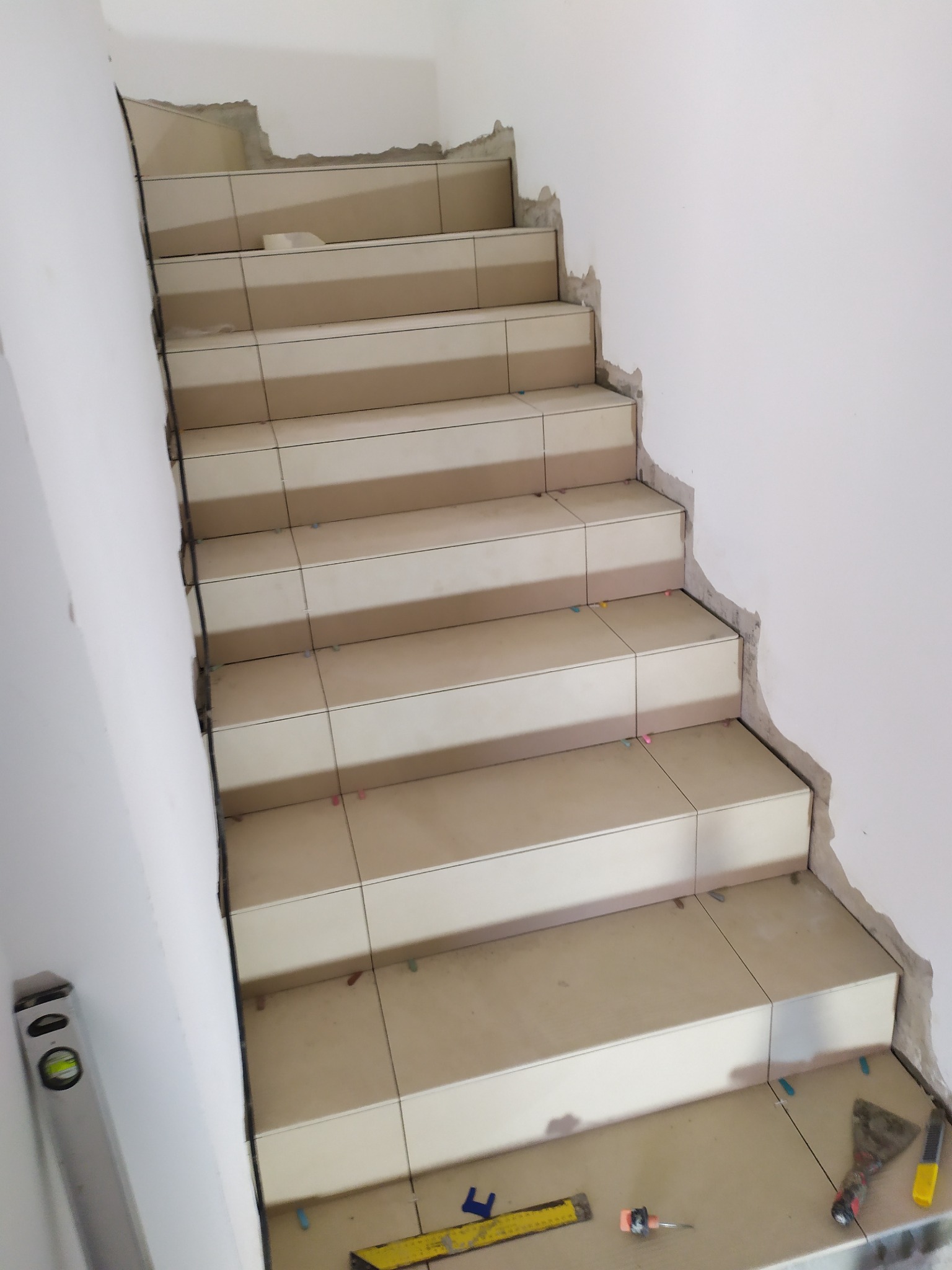 Schody w trakcie wykańczania, z jasnymi płytkami i widocznymi narzędziami glazurniczymi, poziomica przy ścianie, nierówne krawędzie ścian wymagające wykończenia.