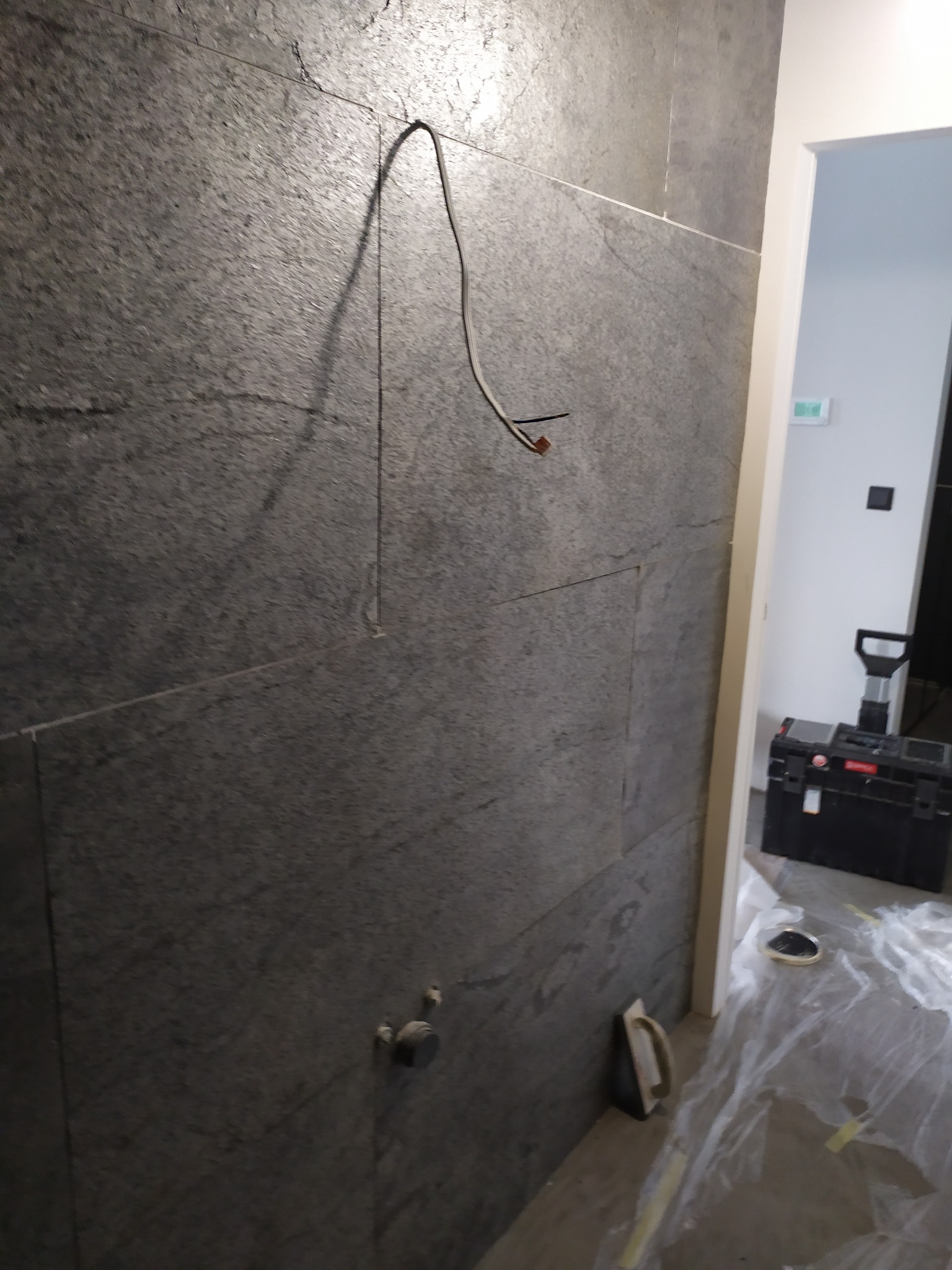 Ściana wyłożona szarymi płytkami imitującymi kamień, widoczne wystające kable, narzędzia glazurnicze i skrzynka narzędziowa w tle, zabezpieczona folią podłoga.