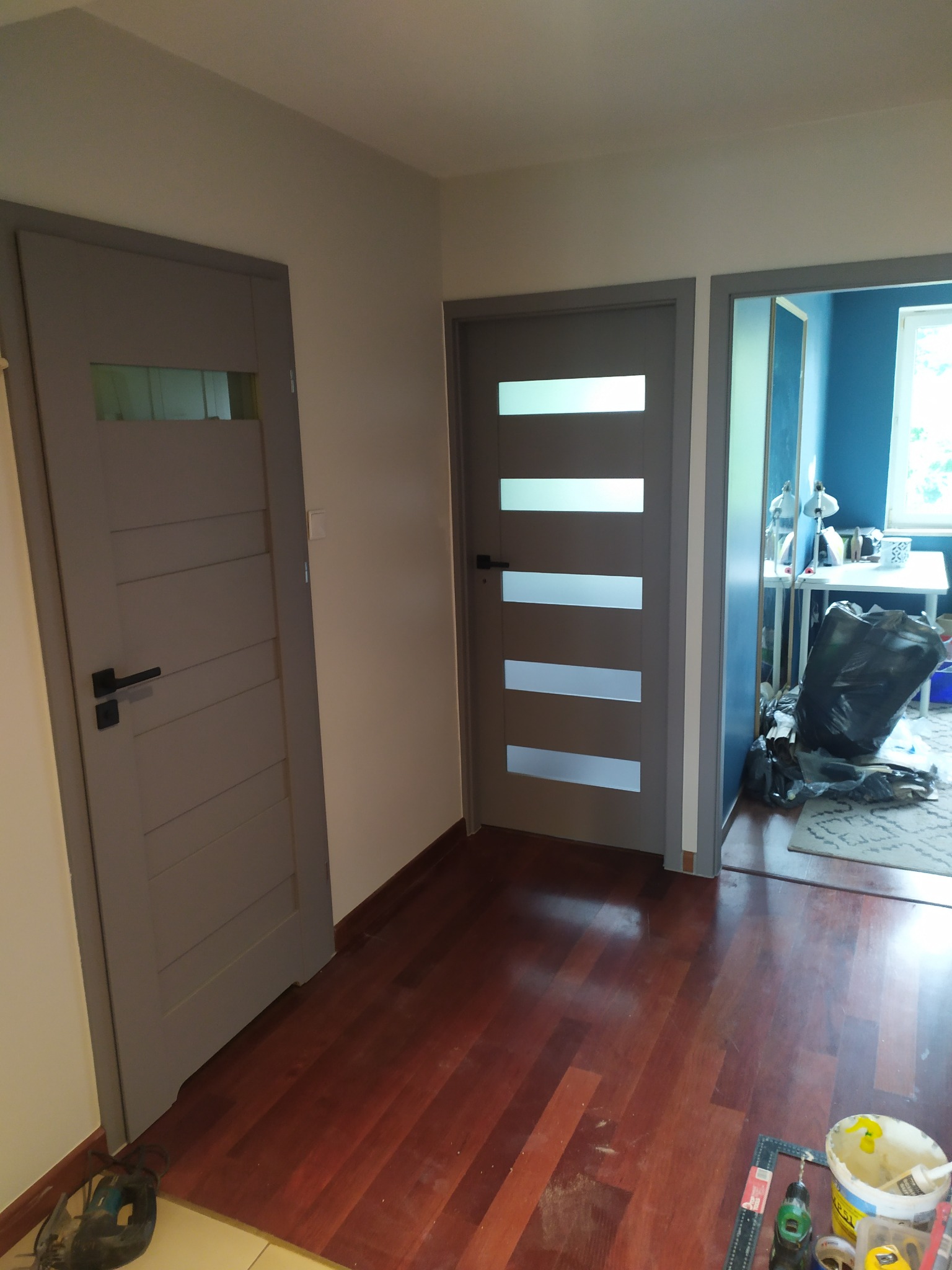 Dwa szare, nowoczesne drzwi z przeszkleniami, zamontowane w odnowionym wnętrzu z ciemną, drewnianą podłogą; widoczne narzędzia i materiały malarskie na podłodze.