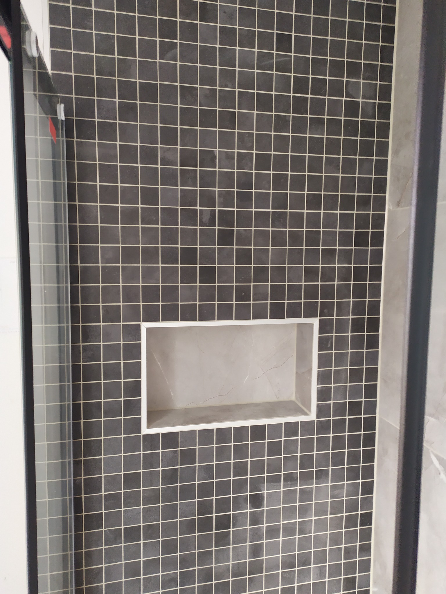 Ściana kabiny prysznicowej wyłożona ciemnymi, kwadratowymi płytkami z jasną fugą; w ścianie wnęka wykończona marmurem.