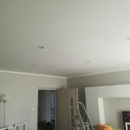 Jasne wnętrze po odświeżeniu: szare ściany, biały sufit z zamontowanymi punktowymi lampami, biała listwa przysufitowa, drabina oparta o ścianę, widoczny fragment telewizora i lampy podłogowej.