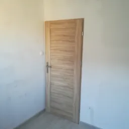 Nowe, dębowe drzwi wewnętrzne z poziomym usłojeniem, zamontowane w odświeżonym pokoju z pomalowanymi na biało ścianami i widocznym gniazdkiem elektrycznym.