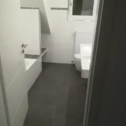 Wykończona łazienka w odcieniach bieli i szarości, z wanną, umywalką, toaletą i oknem, widok zza uchylonych drzwi.