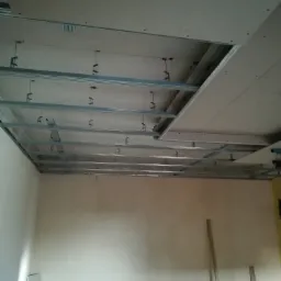 Montaż sufitu podwieszanego z płyt kartonowo-gipsowych, widoczna metalowa konstrukcja nośna i częściowo zamontowane płyty, ściany w stanie surowym.