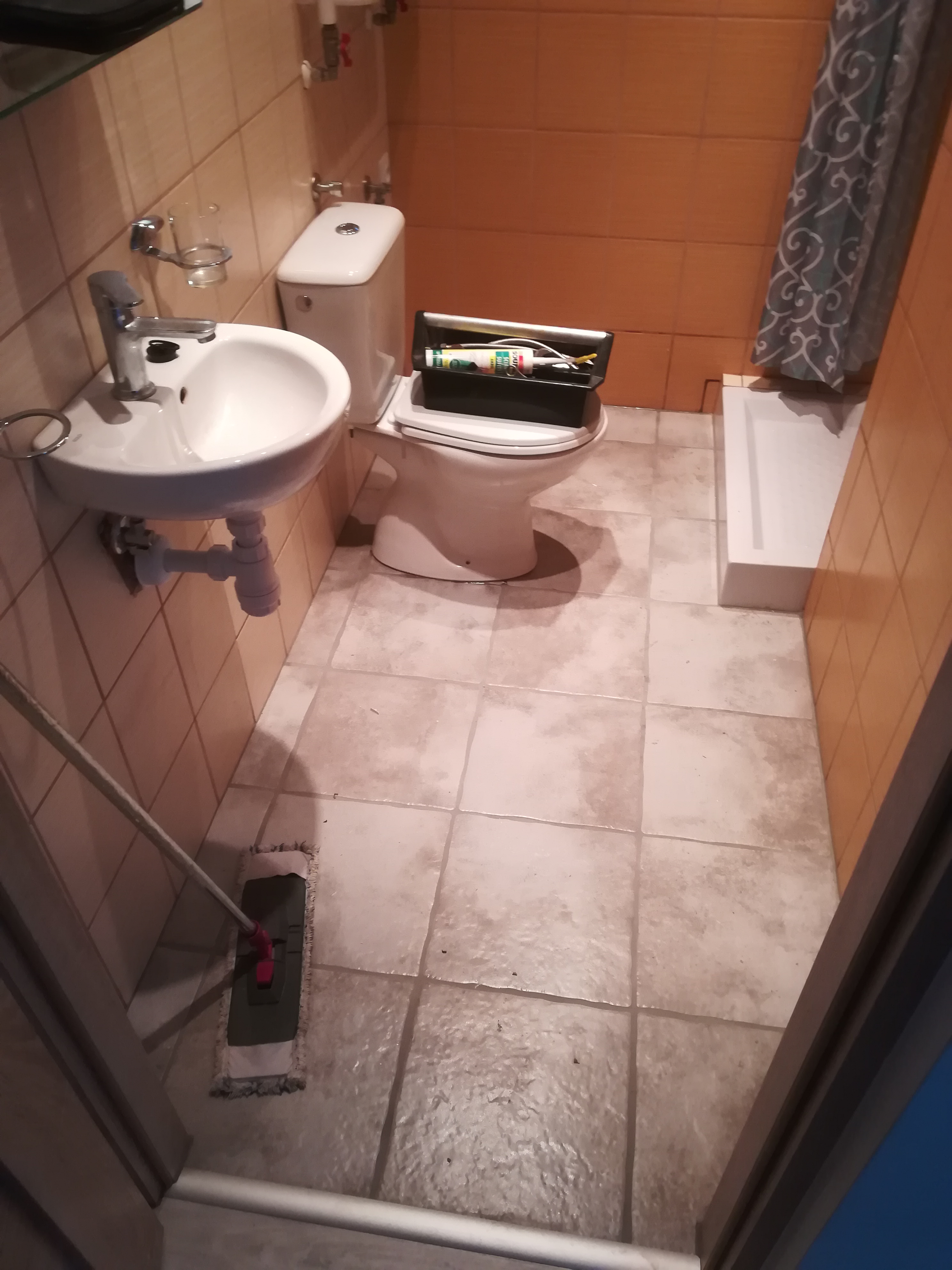 Wnętrze małej łazienki z umywalką, toaletą z otwartym pojemnikiem na narzędzia i mopem do podłogi, ukazujące etap po zakończeniu prac, z widocznymi płytkami na ścianach i podłodze.