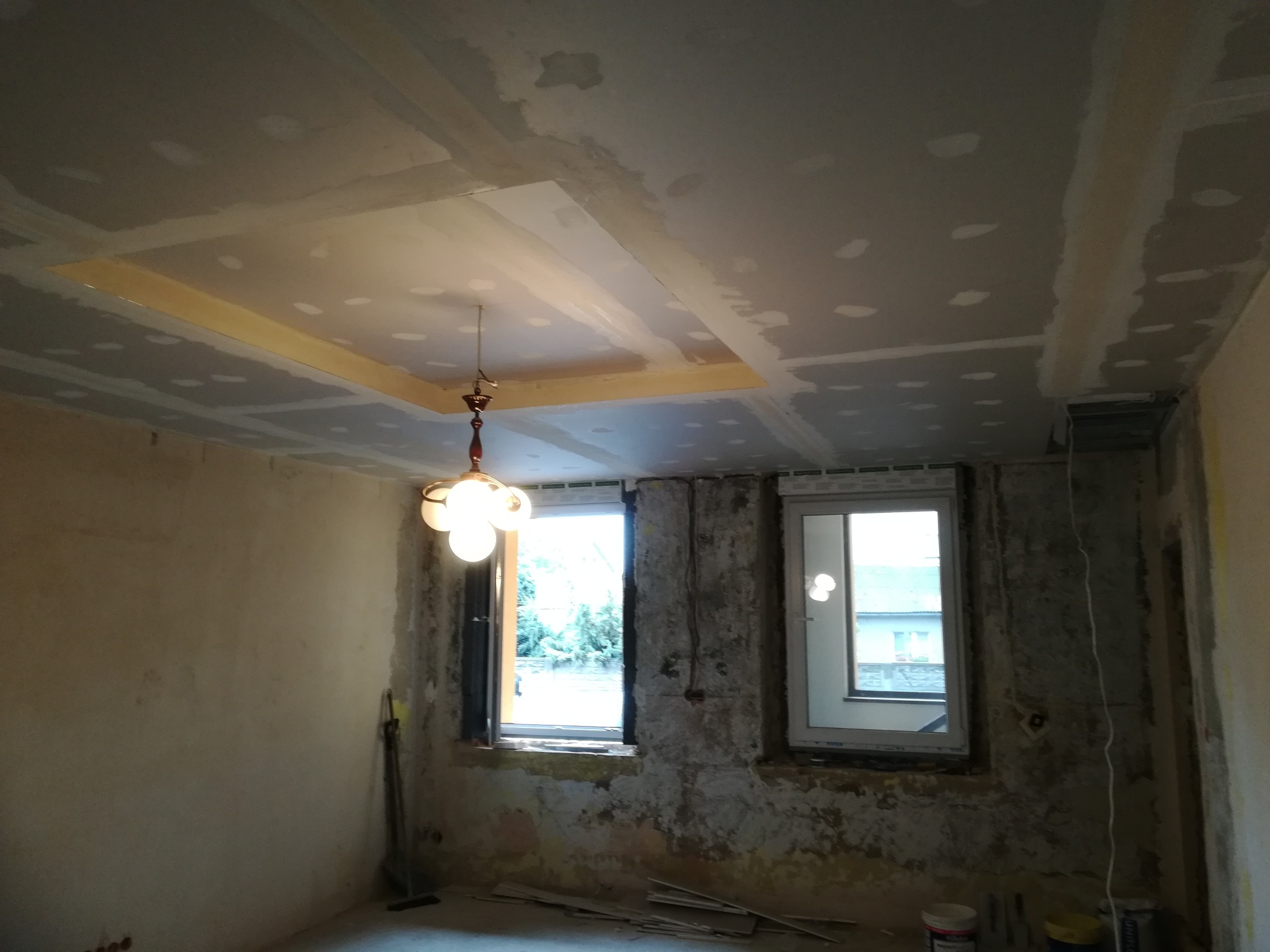 Wnętrze w trakcie prac przygotowawczych: sufit z płyt kartonowo-gipsowych z wyciętym otworem i widoczną konstrukcją, ściany częściowo otynkowane, dwa okna z widokiem na zewnątrz, wisząca lampa...