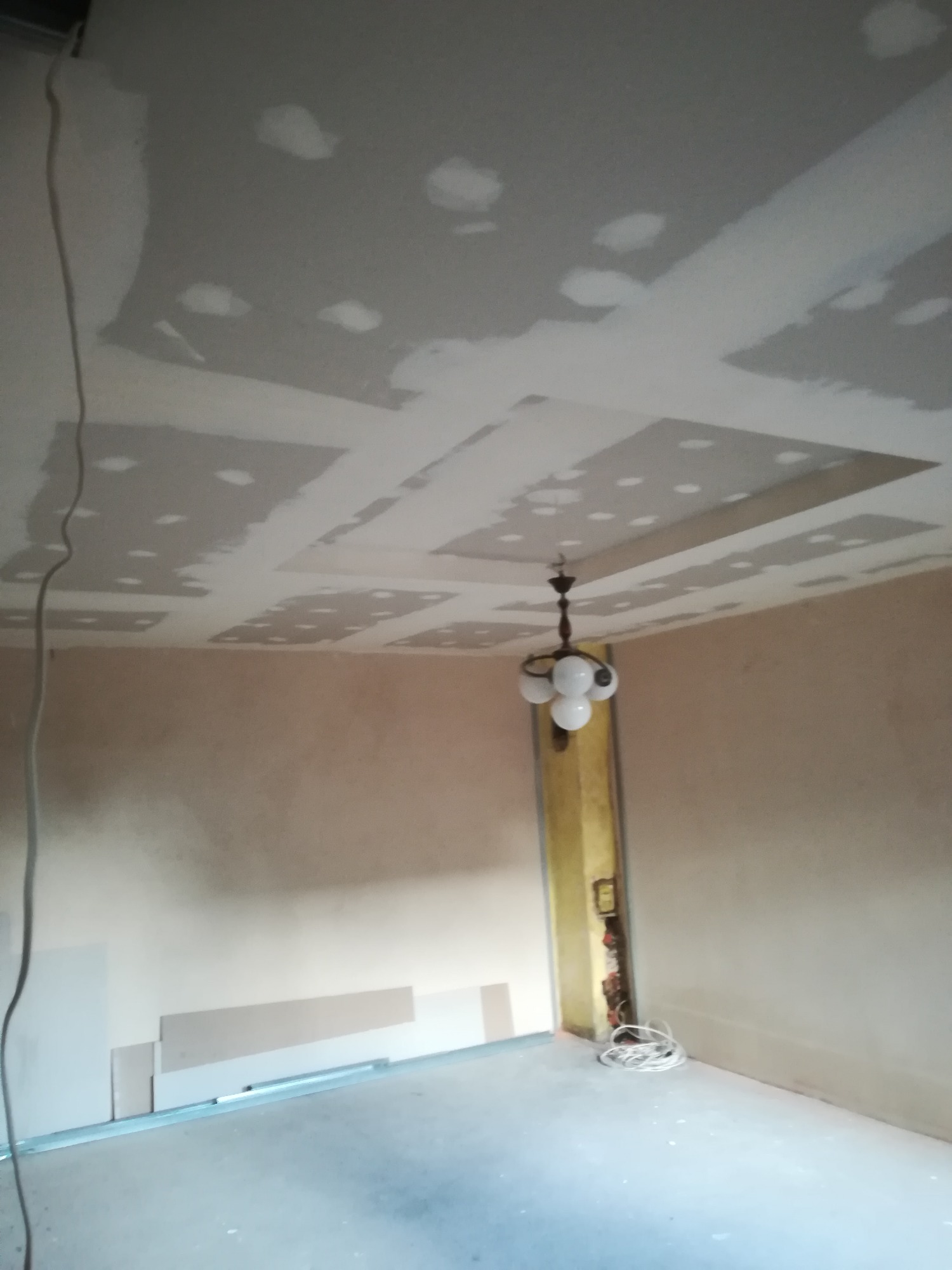 Pokój w trakcie prac wykończeniowych, widoczny sufit z płyt kartonowo-gipsowych z widocznymi łączeniami do szpachlowania,  częściowo przygotowana ściana i zwisający przewód elektryczny.